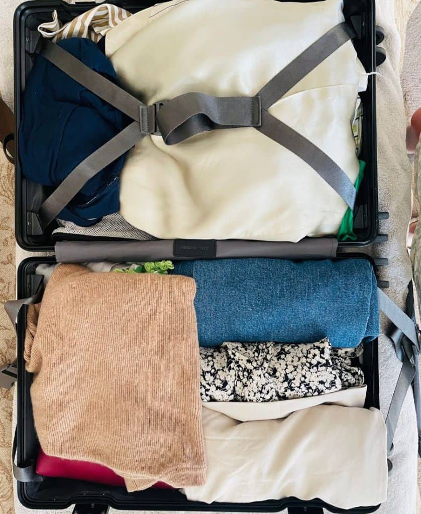 A suitcase is open and filled with clothing of different colors. One side has straps enclosing it.