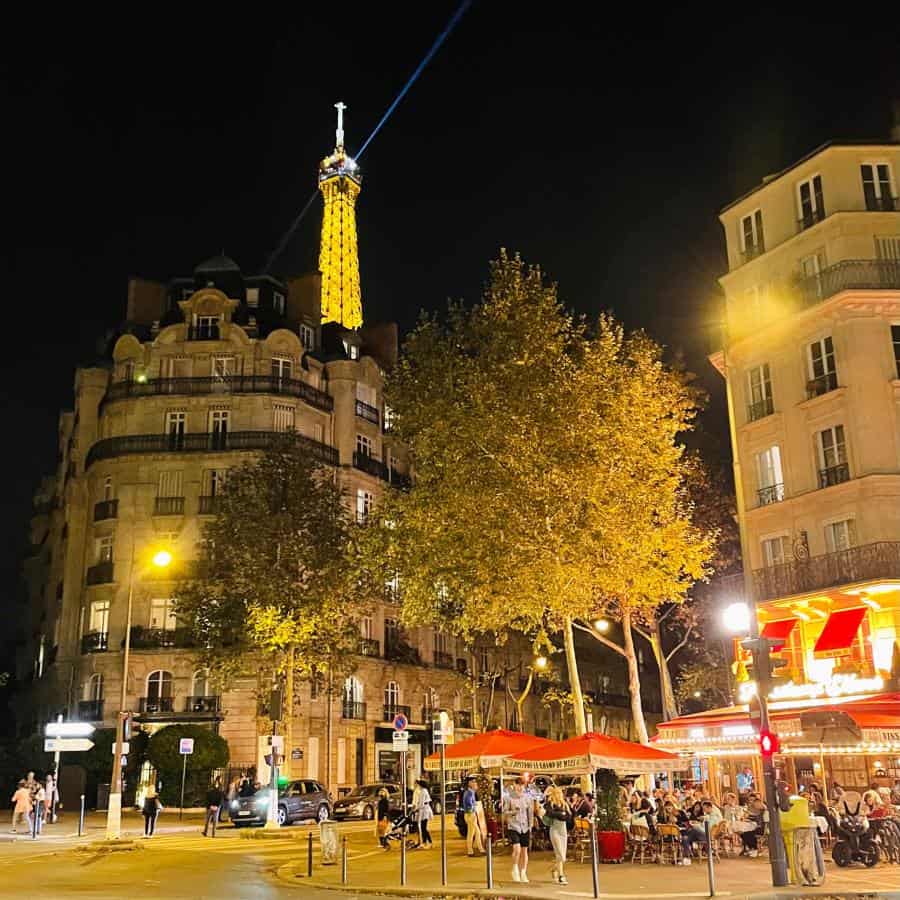A Paris street scene at nighttime with a golden light coming from a cafe with an outdoor terrace with lots of people, and the top of the Eiffel Tower visible above a building with traditional Haussmann architecture