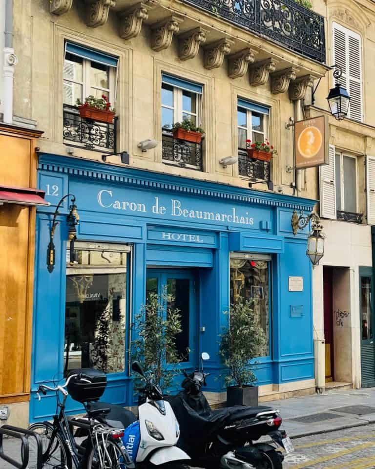 A colorful and charming exterior to a small hotel in Paris. The entrance is flanked in blue trim and lanterns, and above that are three traditional windows with red flowers in flower boxes.