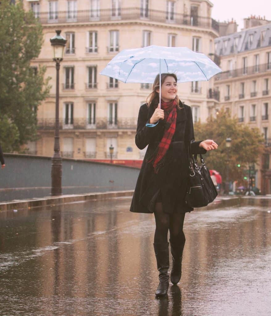 A woman walks in Paris in the rain wearing black boots, a black coat and red scarf. She is holding a blue umbrella and smiling. Haussmann-style buildings are visible in the background.