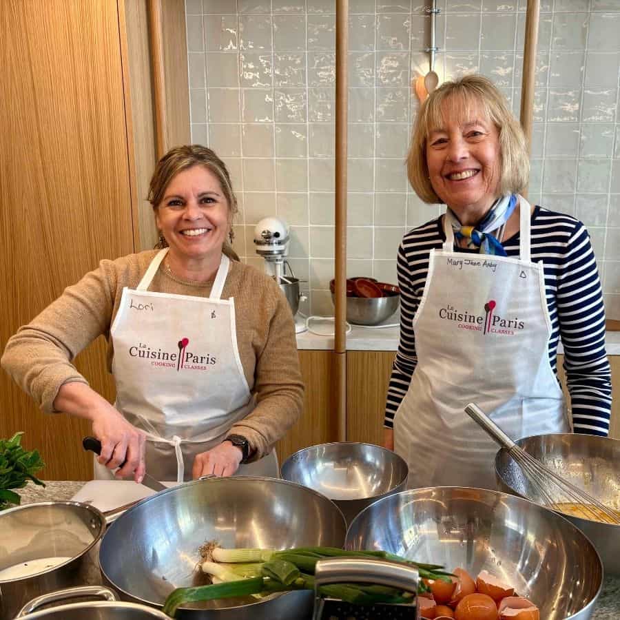 Two women wearing white aprons prepare food with a series of large bowls of ingredients in front of them.