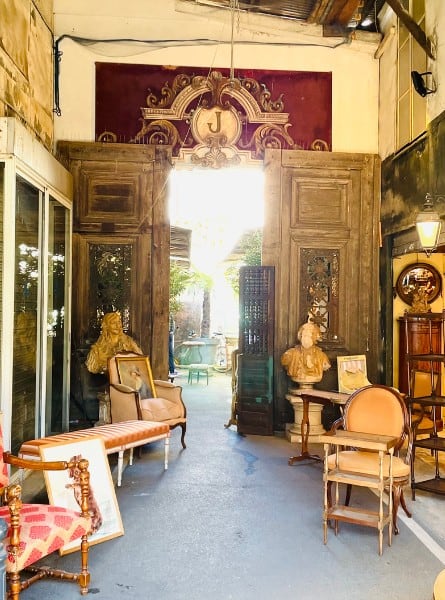 The Paris Les Puces, the largest Antique Market in the world. Pictured are an array of antique furniture and a lantern. An open door is flanked by tall wooden doors. And an assortment of curio and books.