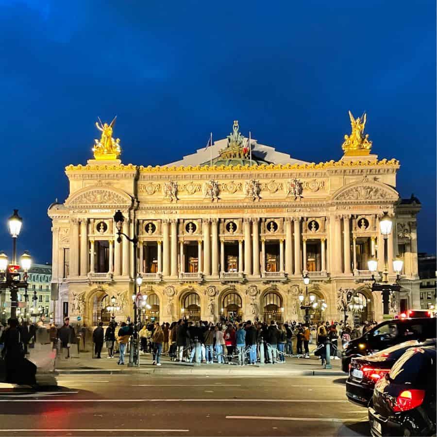 The exterior of the Paris Palais Garnier at Night. The beautiful art nouveau exterior with columns and statues. A crowd of people are standing in front and some cars are visible on the right of the screen.
