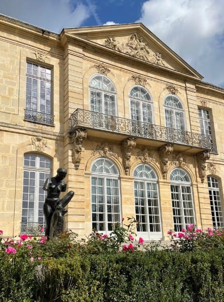 The exterior of the Rodin Museum in Paris with a decorative stone facade. In the foreground is a garden with pink roses and a sculpture of a woman.