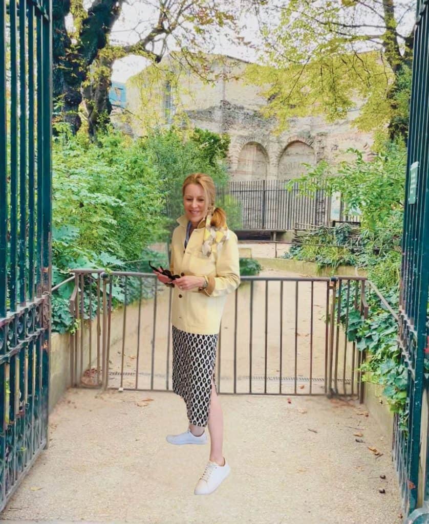 A woman stands in front of a park gate in Paris. She is wearing a black and white patterned skirt, white runners and a yellow barn jacket. She is holding sunglasses and has a scarf tied in her hair.