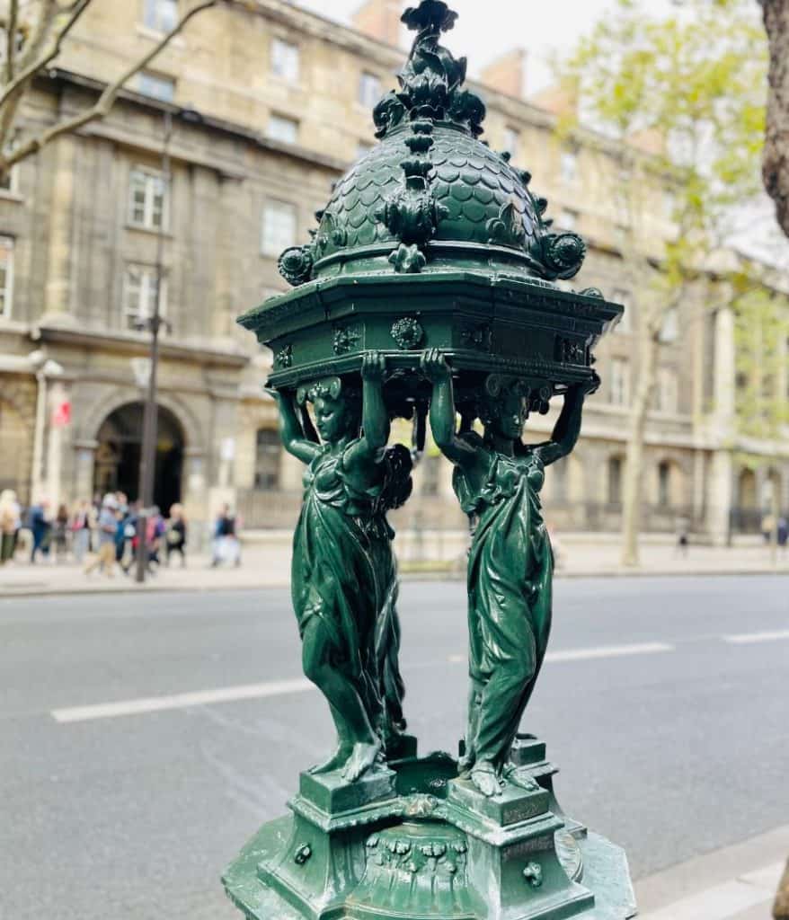 One of the 108 Wallace Fountains in Paris. It is green metal and has a distinctive design featuring four caryatids (sculptures of women) supporting a canopy.