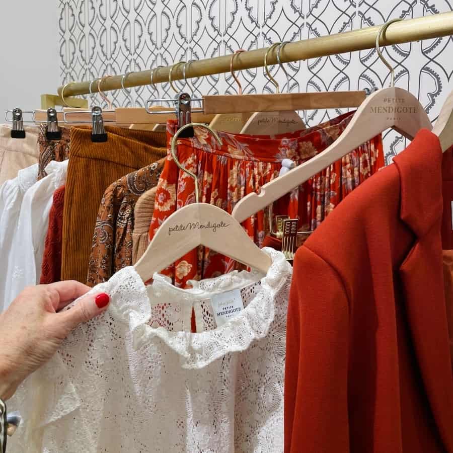 A woman's hand holds a white blouse next to blazers and skirts in fall colours at Petite Mendigot, an affordable French brand in Paris
