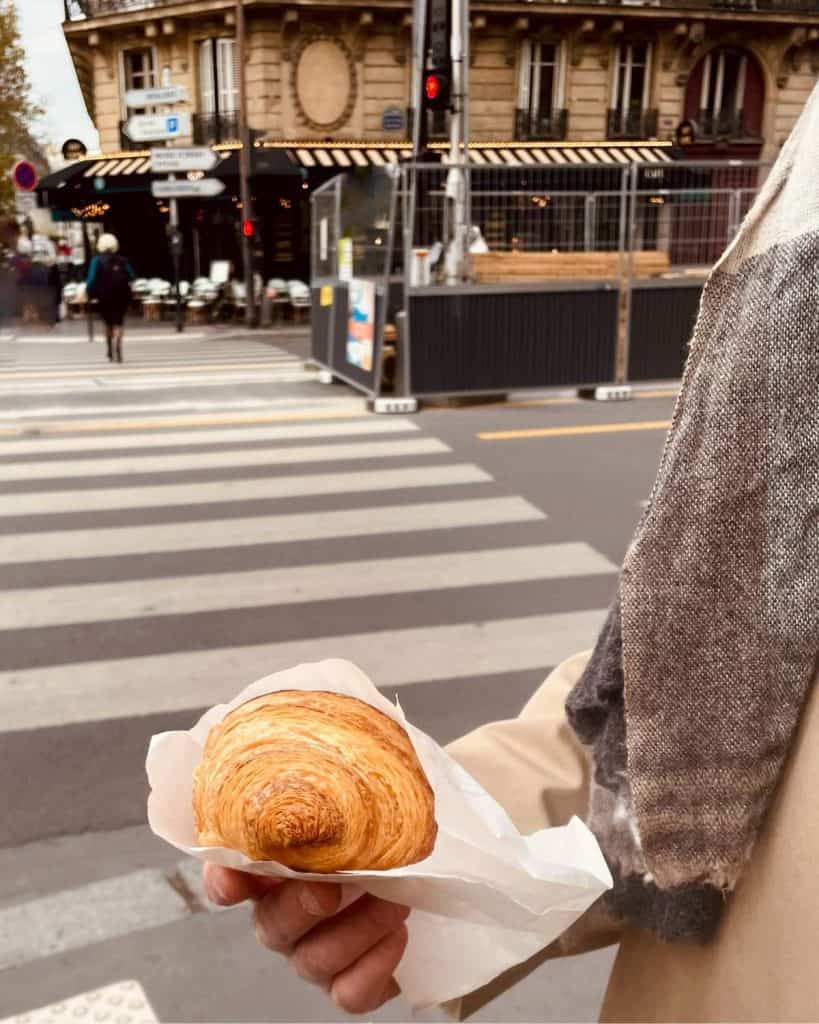 A man holds a butter croissant on a street in Paris. Only his arm and trench coat are visible.