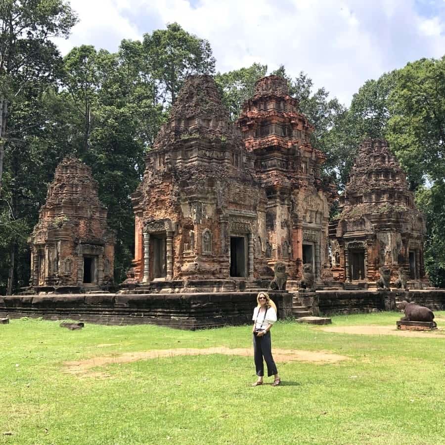 Susan Heinrich stands on from of an ancient temple complex known as Preah Ko, in Cambodia. Four spires are visible on a platform as well as trees in the distance.