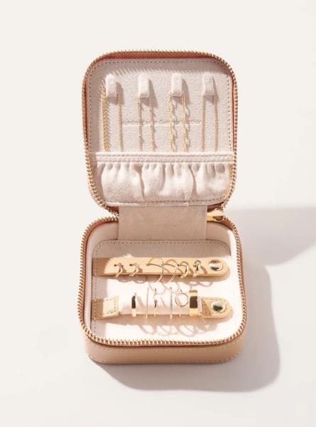 A square travel jewelry case make an ideal gift for a travel lover. Pictured open with necklaces, earrings and rings. Beige with gold hardware. Made by Quince.