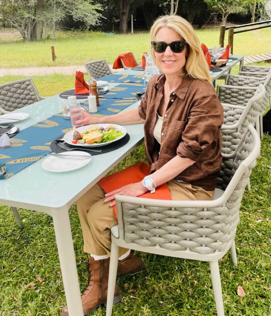 Susan Heinrich sits at a lunch outdoors at Leroo la Tau safari lodge, Botswana wearing an African safari outfit, brown linen short, tan pants and brown safari-style boots.