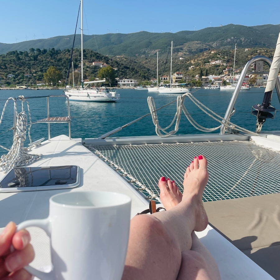 Susan sits with a cup of coffee on a sailboat in the Saronic Greek island. Only her legs are visible, with sailboats in the distance