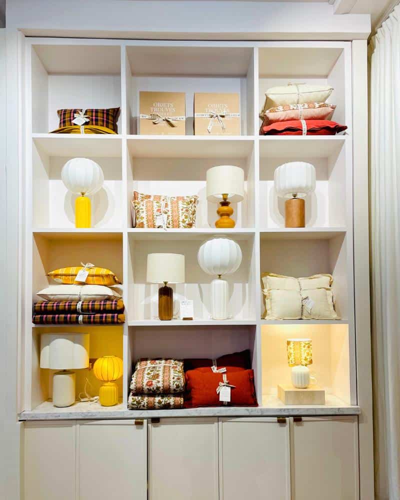 A selection of the new Interiors Collection by Sézanes, Les Composantes. Pictures are lamps and linens set in a white shelving unit. Colors are primarily yellow and red.