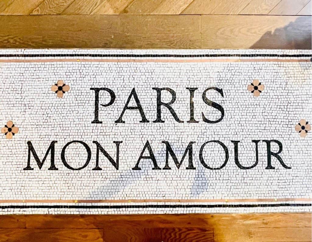 Decorative floor tiles are inlaid in a wood floor at a Sézane store in Paris, France. The tiles are white and the words "Paris Mon Amour" are spelled in black.