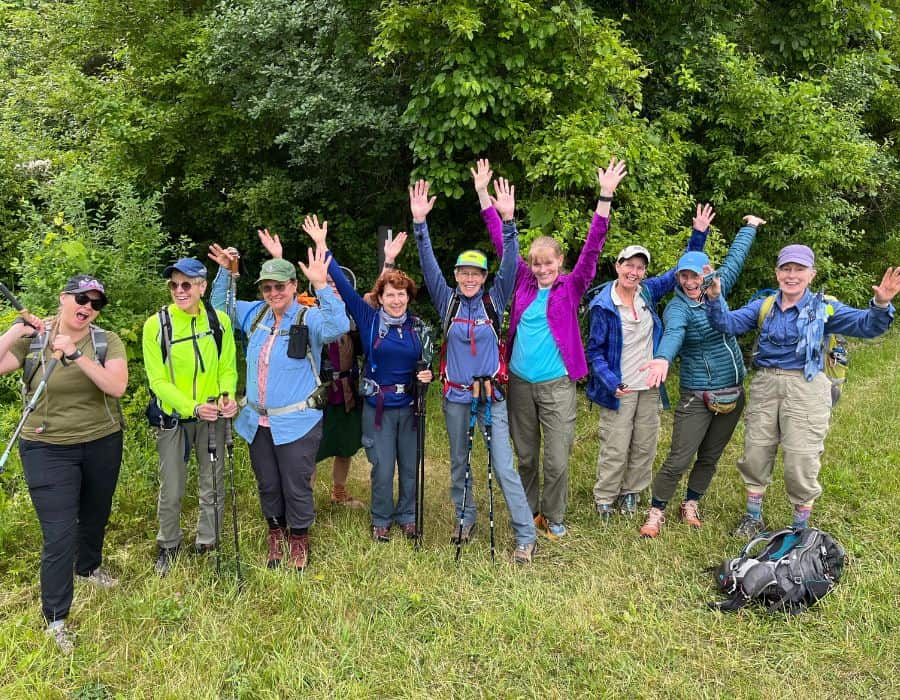 Ten women stand on front of a forest area, on a trail. They are on a slackpacking trip with the company, Adventures in Good Company, in Massachusetts.