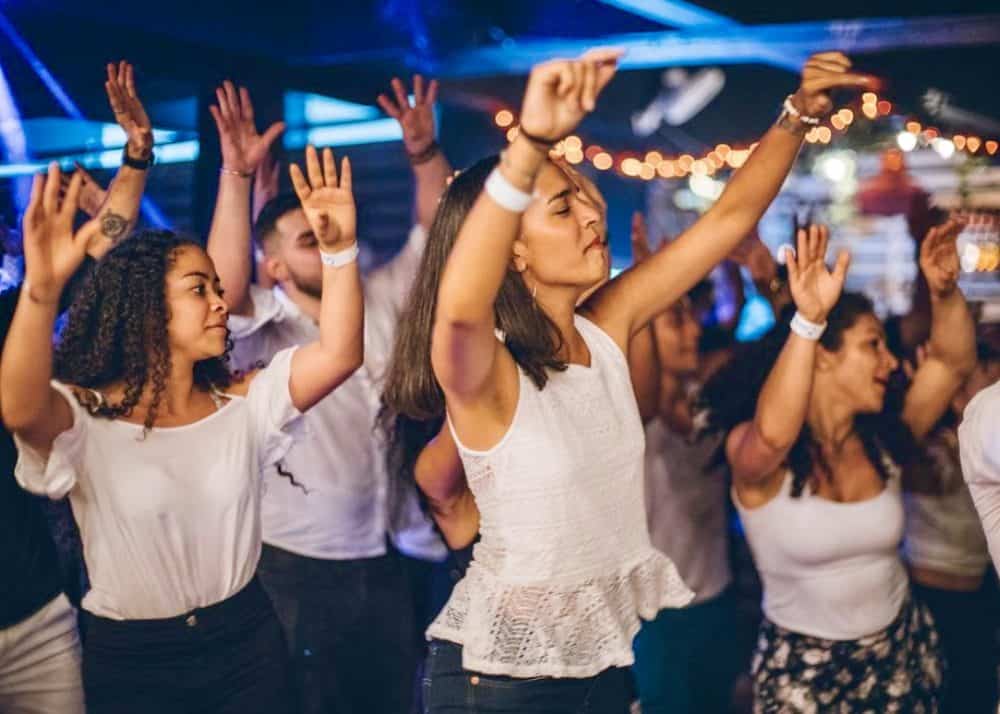 A group of mostly young women have their hands in the air during a dance class. They are all wearing white tops.
