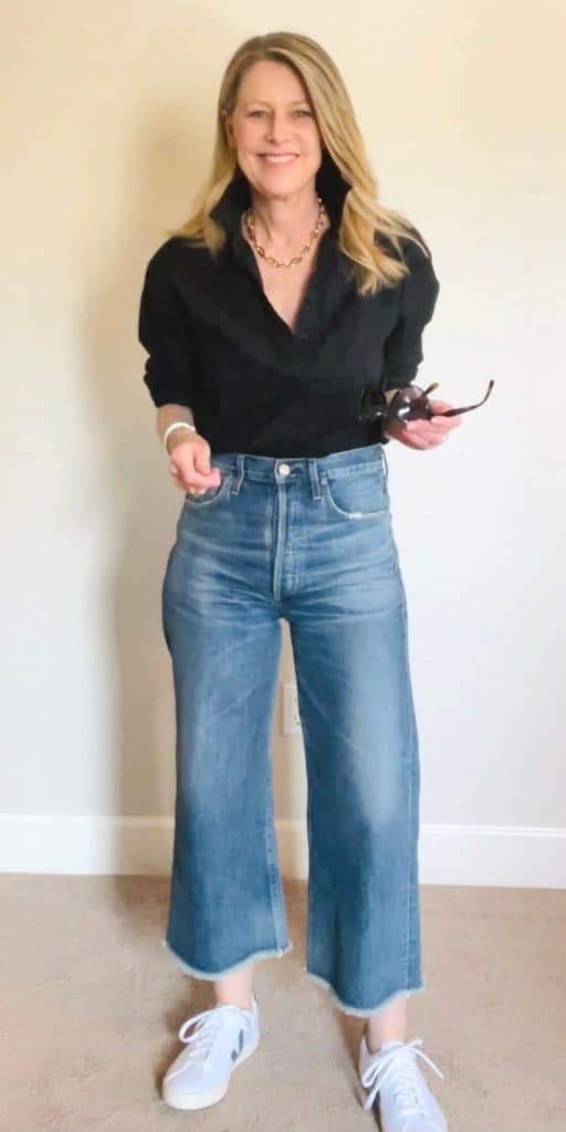 Susan in cropped jeans and a black shirt, and holding sunglasses