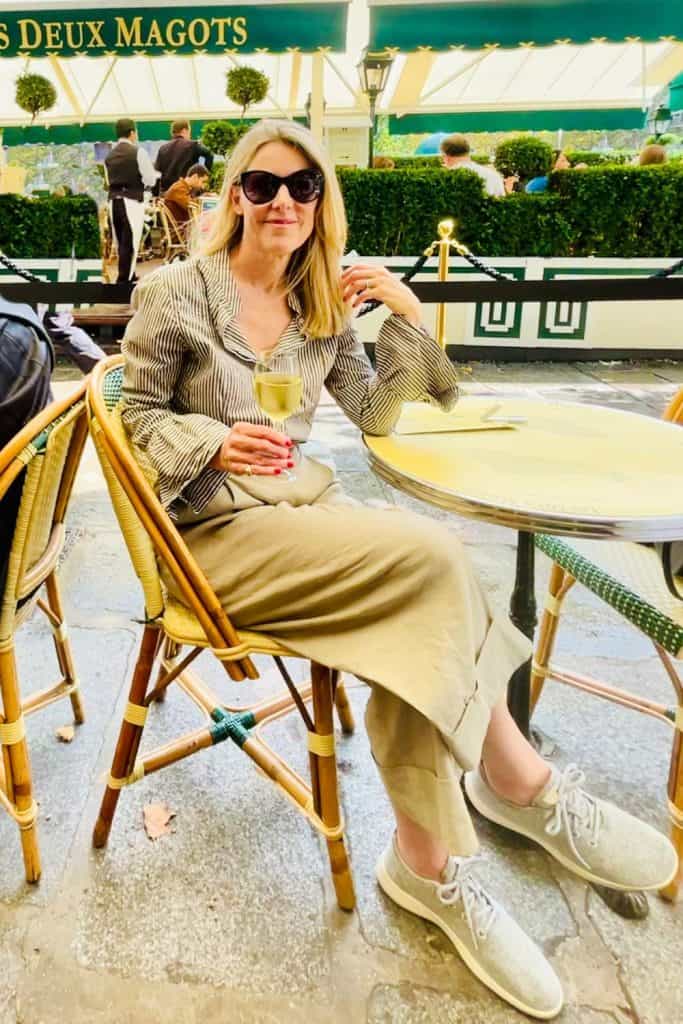 A woman sits in a Paris café holding a glass of white wine. She is wearing tan trousers, a striped button-up shirt, sneakers and sunglasses.