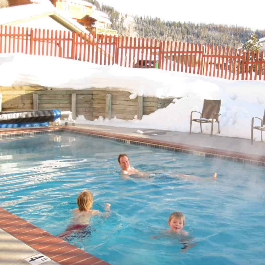 A man and two children swim in an outdoor pool in winter with snow around the exterior.
