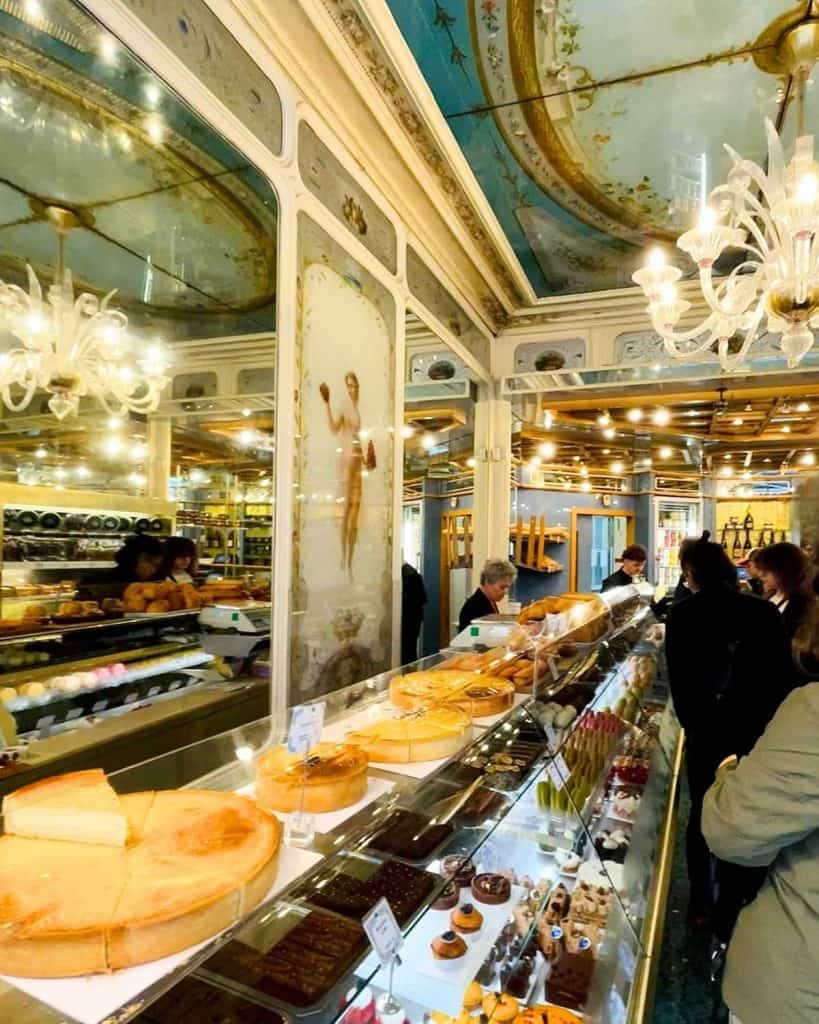 A glass case inside an ornate Paris patisserie is filled with colorful pastries and cakes. A chandelier is partially visible.