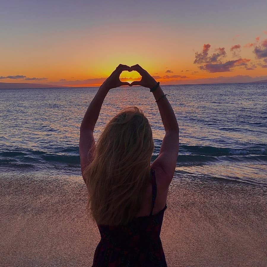 A woman holds up her hands to make a heart around the sunset at Kaanapali Beach in Maui