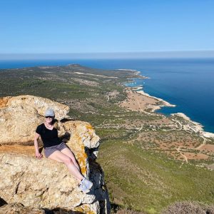 Susan Heinrich sits on a rock with the Cyprus coastline and turquoise sea beyond. The famous blue lagoon can just be seen. She is wearing shorts and a tee and a baseball hat and is smiling.