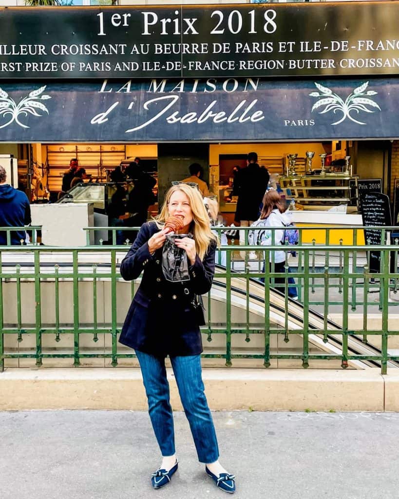 Susan Heinrich stands in from the of Maison d'Isablle bakery in Paris biting a croissant.