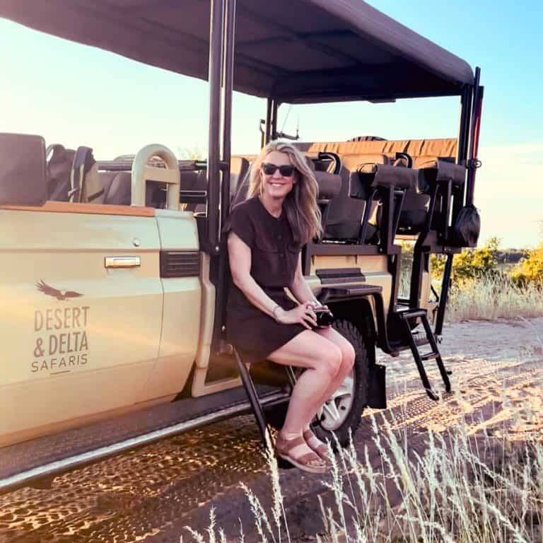 Susan Heinrich sits on the side of a safari jeep in Botswana Africa wearing a brown safari outfit, a utility safari-style shirt dress and sandals.