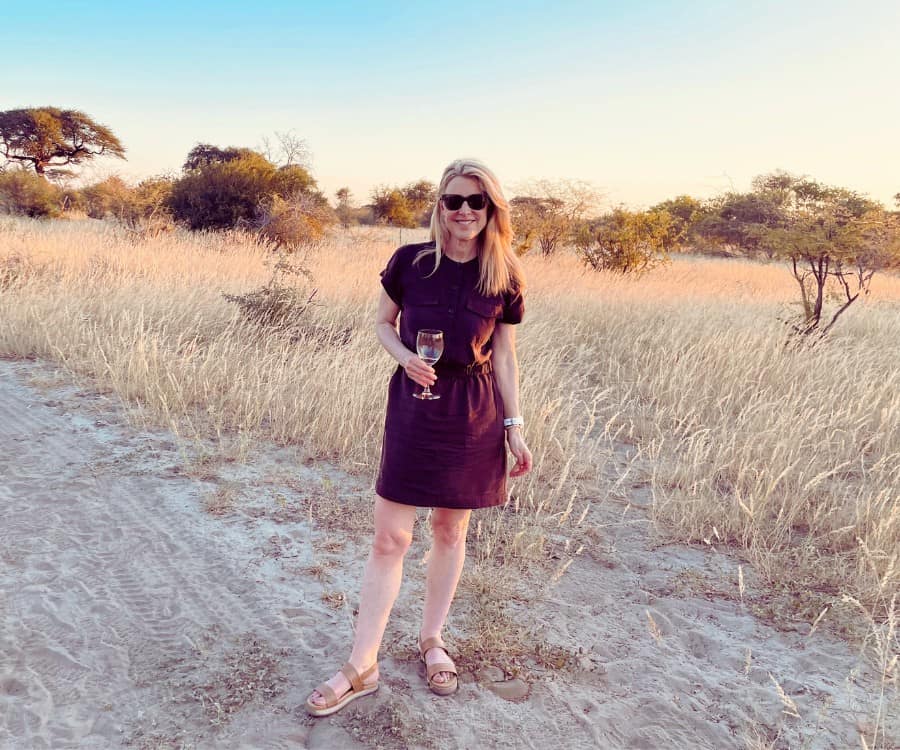 Susan Heinrich wears a brown utility dress and holds a glass of white wine in an open grass area of Botswana's Makgadikgadi Pans National Park.