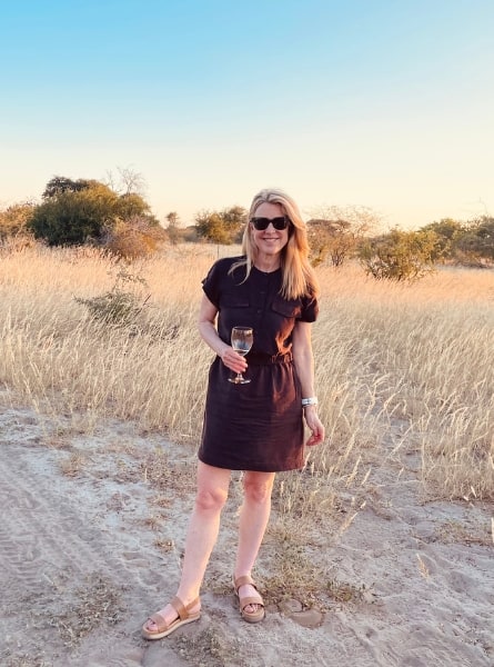 Susan Heinrich stands in an open grassy landscape in Botswana holding a glass of white wine. She is on safari enjoying the traditional sundowner drink, with the sun setting behind her.