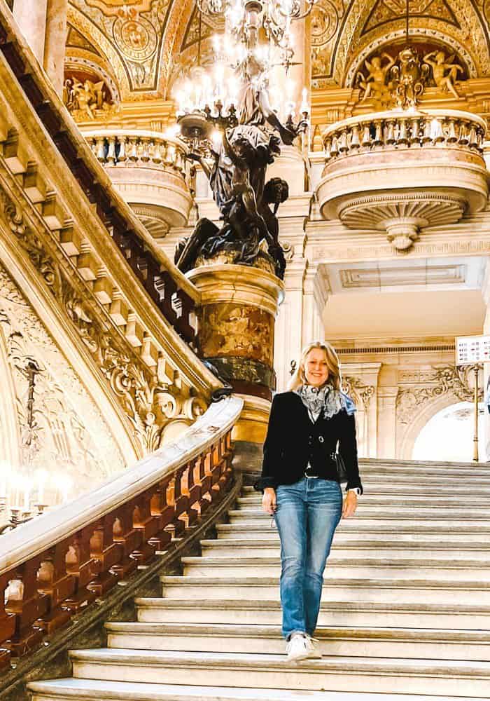 Susan Heinrich stands on the steps of the Palais Garnier Opera House in Paris wearing jeans a classic black blazer and chic sneakers by Veja.