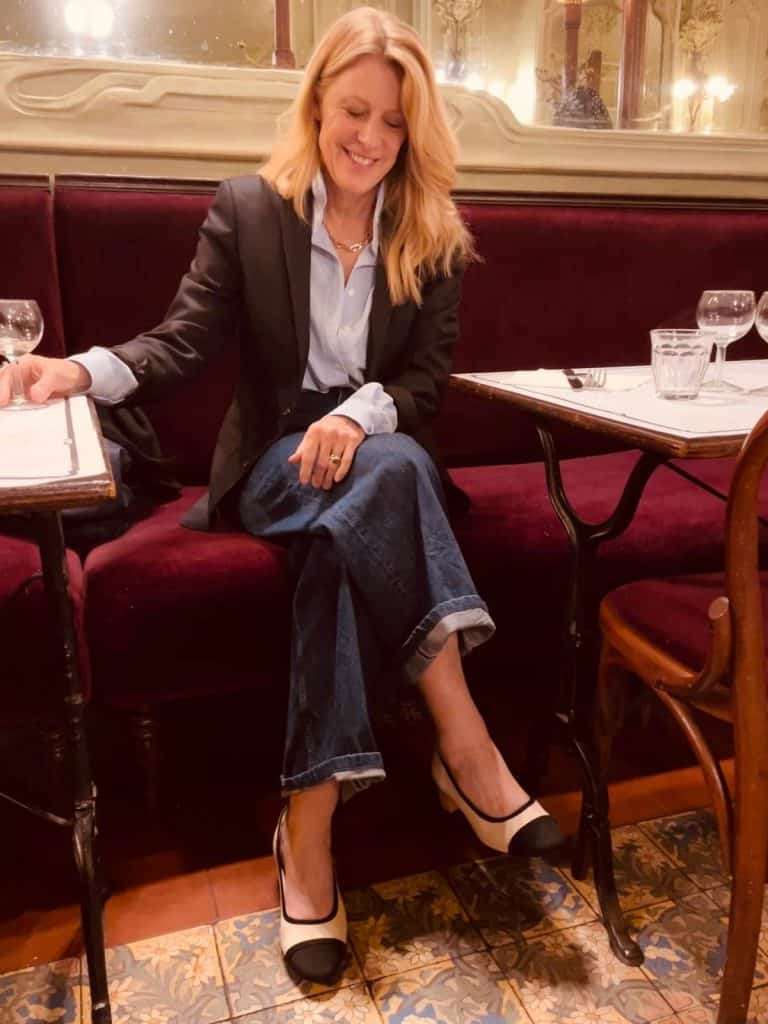 Susan Heinrich sits in Paris Bouillon restaurant on a velvet banquette with a large mirror behind her and lights reflected. The floor is a traditional tile. She is wearing a black blazer, jeans, a blue button-up and block-heel shoes by Vivaia, cream with a black toe-cap.