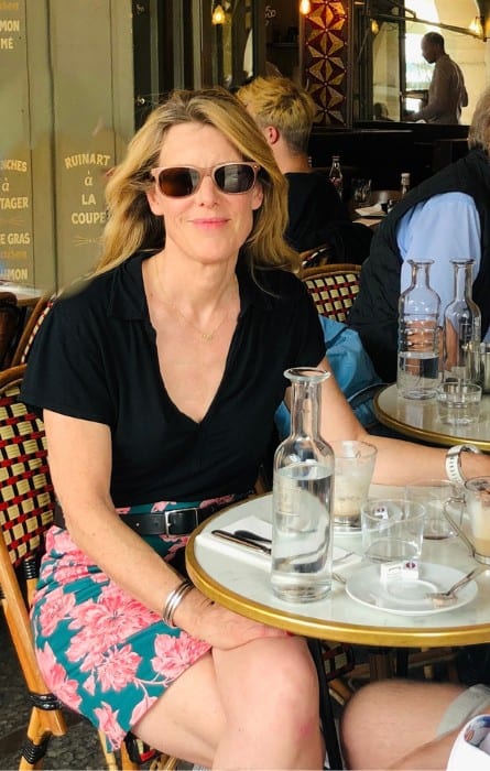 Susan Heinrich sits in a Paris cafe wearing a black short-sleeved v-neck top and flowered skirt with a black belt.