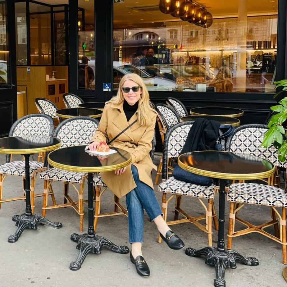 Susan Heinrich sits at a cafe table in Paris. She is wearing a tan trench coat, jeans and black loafers. She has a small black bag accross her. She has a croissant on a table in front of her.