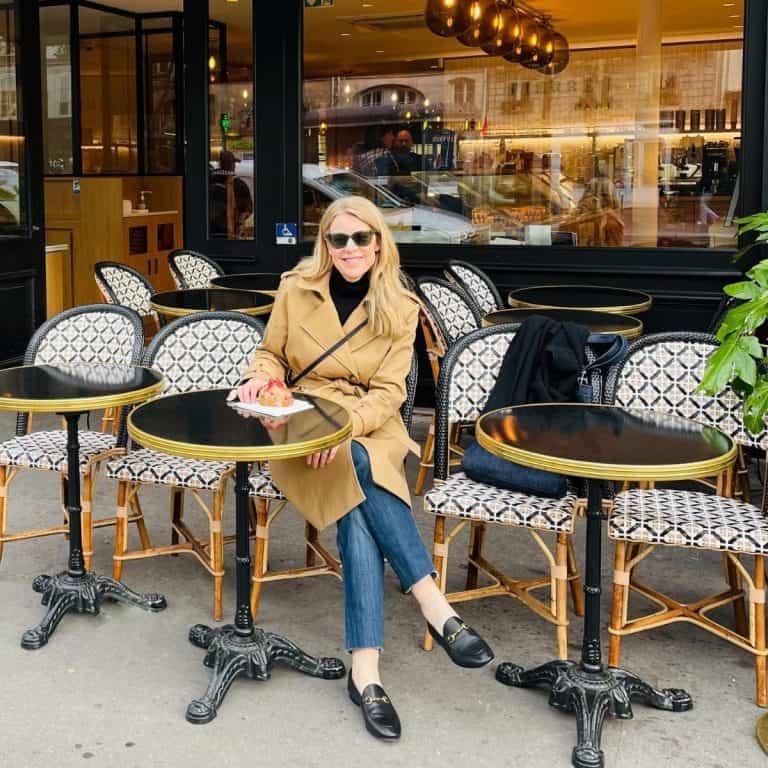 Susan Heinrich sits at a cafe table in Paris. She is wearing a tan trench coat, jeans and black loafers. She has a small black bag accross her. She has a croissant on a table in front of her.