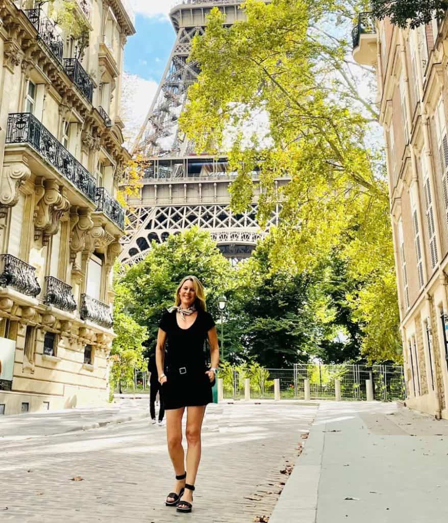 Susan Heinrich stands on a Paris street in late summer with the Eiffel Tower beyond her. The street is lined by beautiful Haussmann-style buildings. She is wearing black shorts and a black tee and scarf and black platform sandals by Franco Sarto.