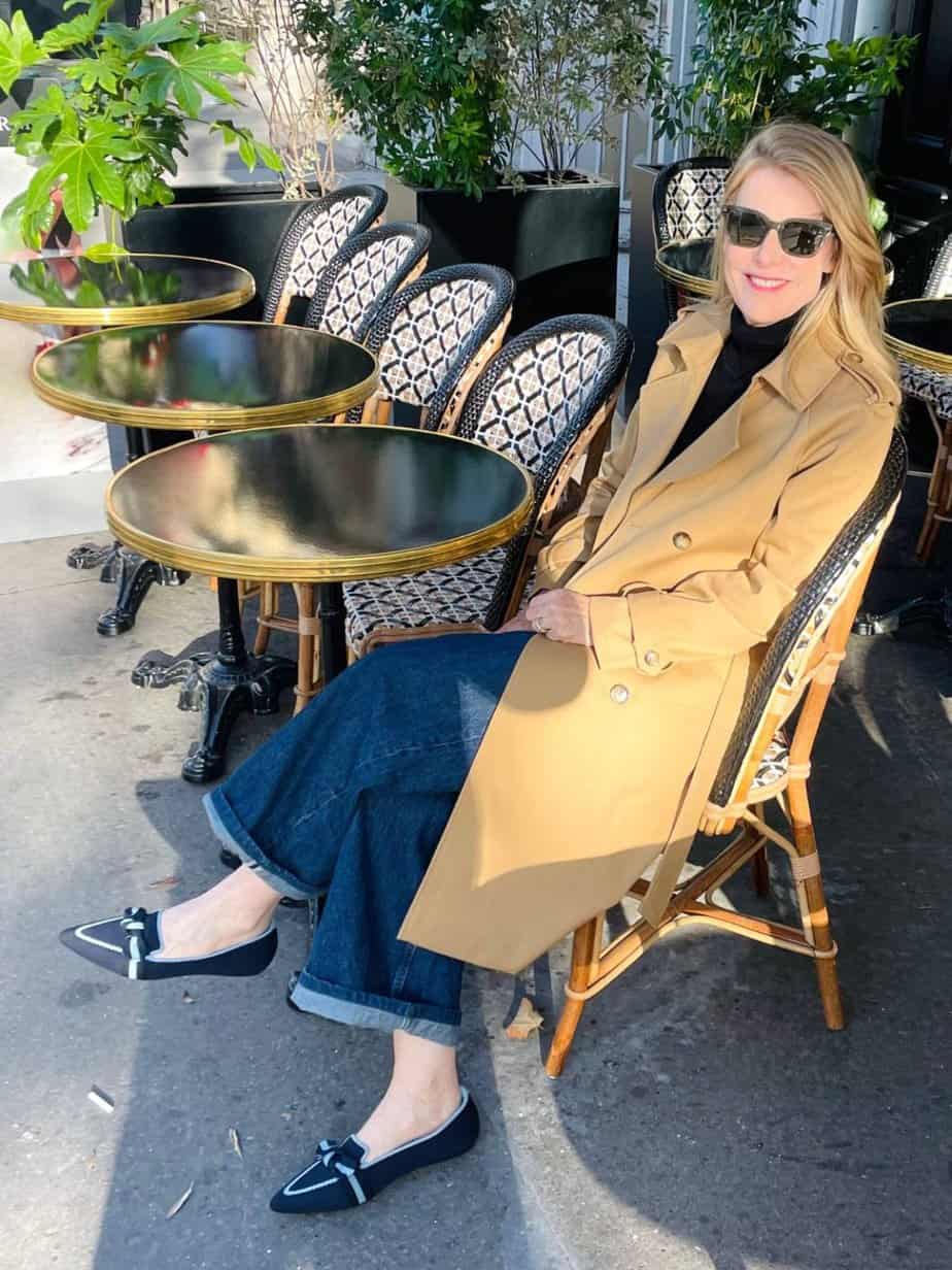 Susan Heinrich sits outdoors at a Paris cafe in fall wearing a trench coat, wide-leg jeans and blue pointed-toe flats.