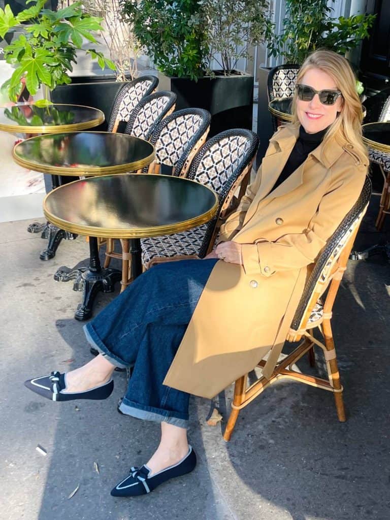 Susan Heinrich sits outdoors at a Paris cafe in fall wearing a trench coat, wide-leg jeans and blue pointed-toe flats.