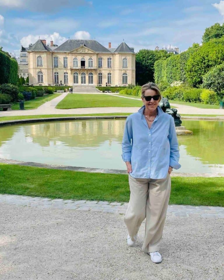 A woman stands in front of a pond and green lawn with a traditional French-style chateau in the distance. She is wearing a blue button-up shirt untucked, and beige trousers, sunglasses and white sneakers.