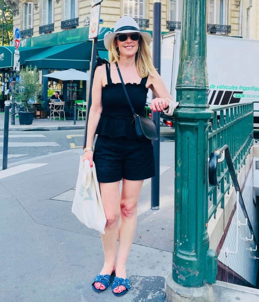 Susan Heinrich stands on a Paris street next to a Metro station entrance in late summer. She is wearing black short and tank top and Paris walking shoes, blue sandal slides by Rothys. She is also wearing a white fedora-style hat and sunglasses.