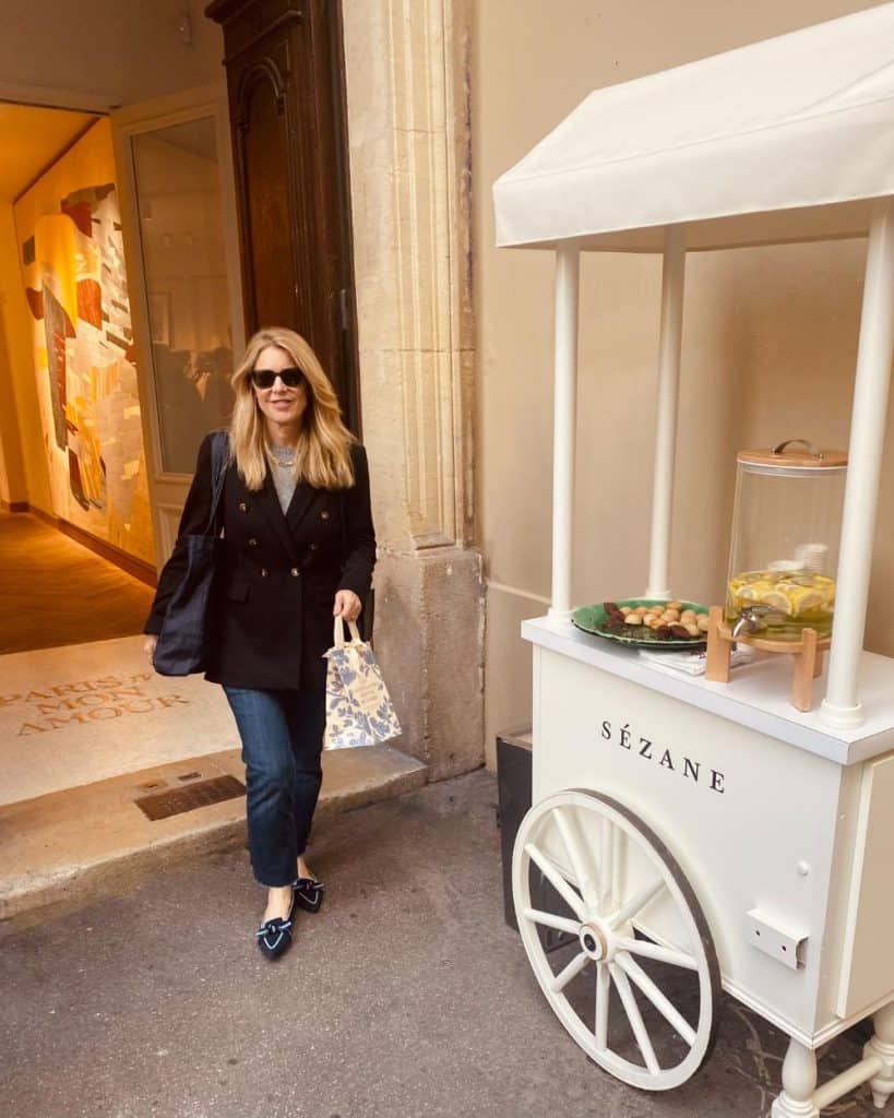 Susan Heinrich in front of the Sézane store in Le Marais. She is holding a small Sézane bag and wearing a black blazer, jeans and loafers.