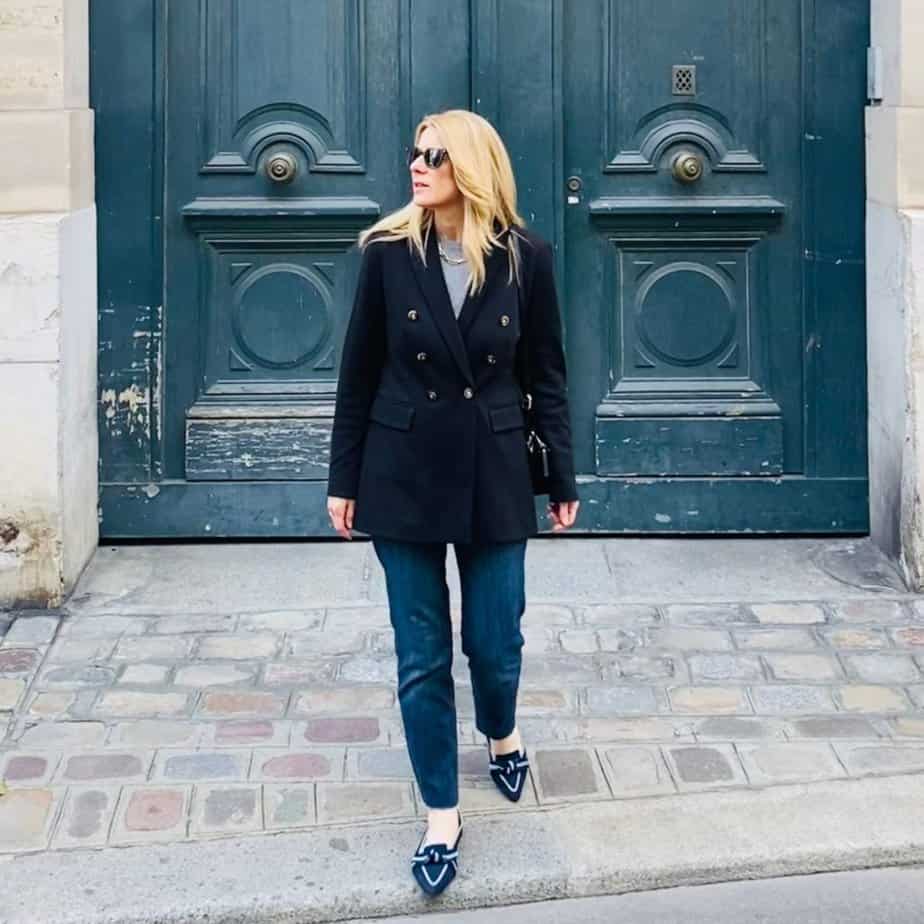 Susan Heinrich stands in front of a large blue door in Paris. She is wearing classic French pieces, denim, a black blazer and ballet flats.