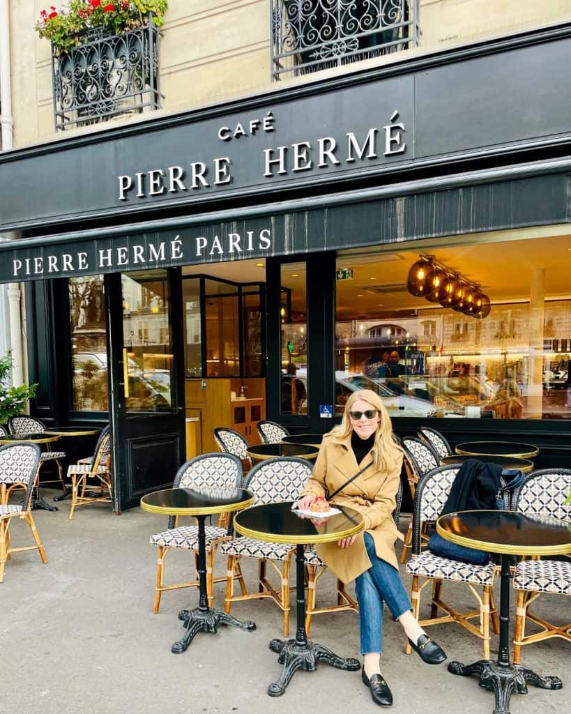 Susan Heinrich sits at a cafe table in front of the Pierre Hermé bakery in Paris. On a plate in front of her is her favorite croissant, the Ispahan. She is wearing a trench coat and jeans.