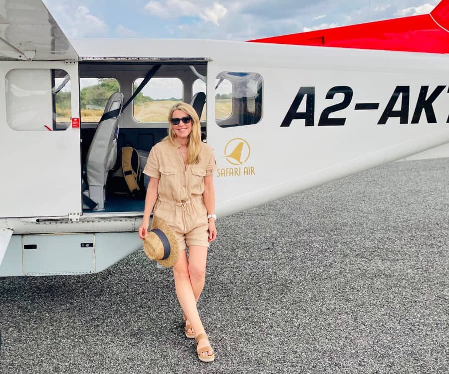 Susan Heinrich leans against the side of a small 6-seater airplane while on safari in Botswana. She wears a beige linen shorts romper and is holding a hat.