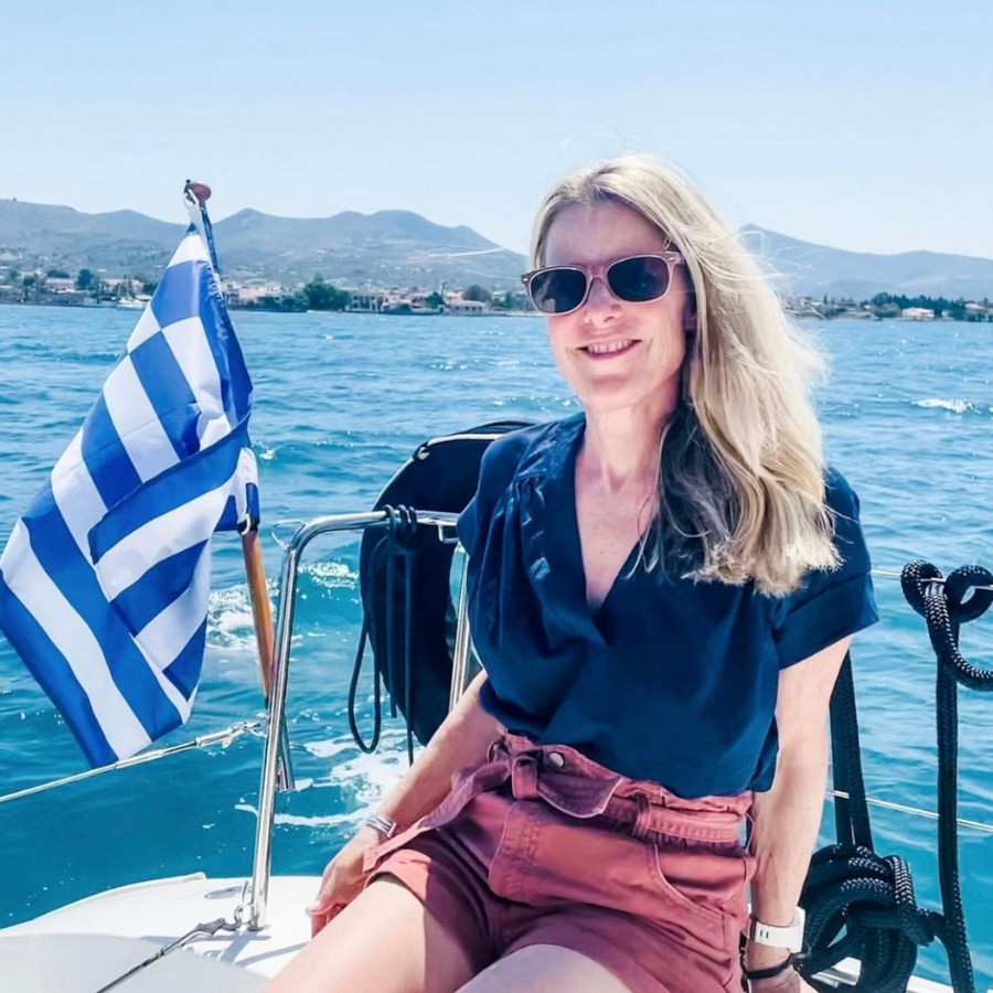 Susan Heinrich sits on a sailboat in the Greek Islands with a Greek flag behind her