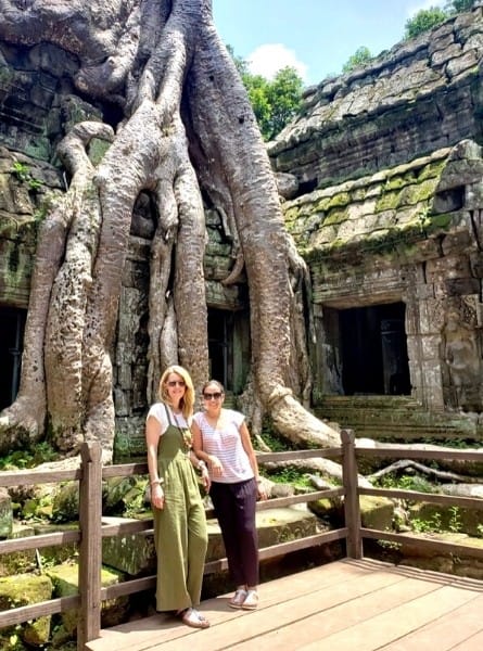 Two women pose giant tree roots which surround a temple at Ta Prohm