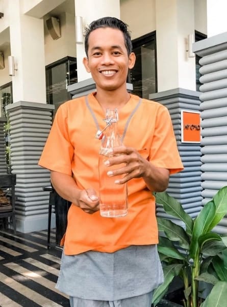 A smiling male staff member named Sophy holds a glass water bottle at a hotel in Siem Reap Cambodia