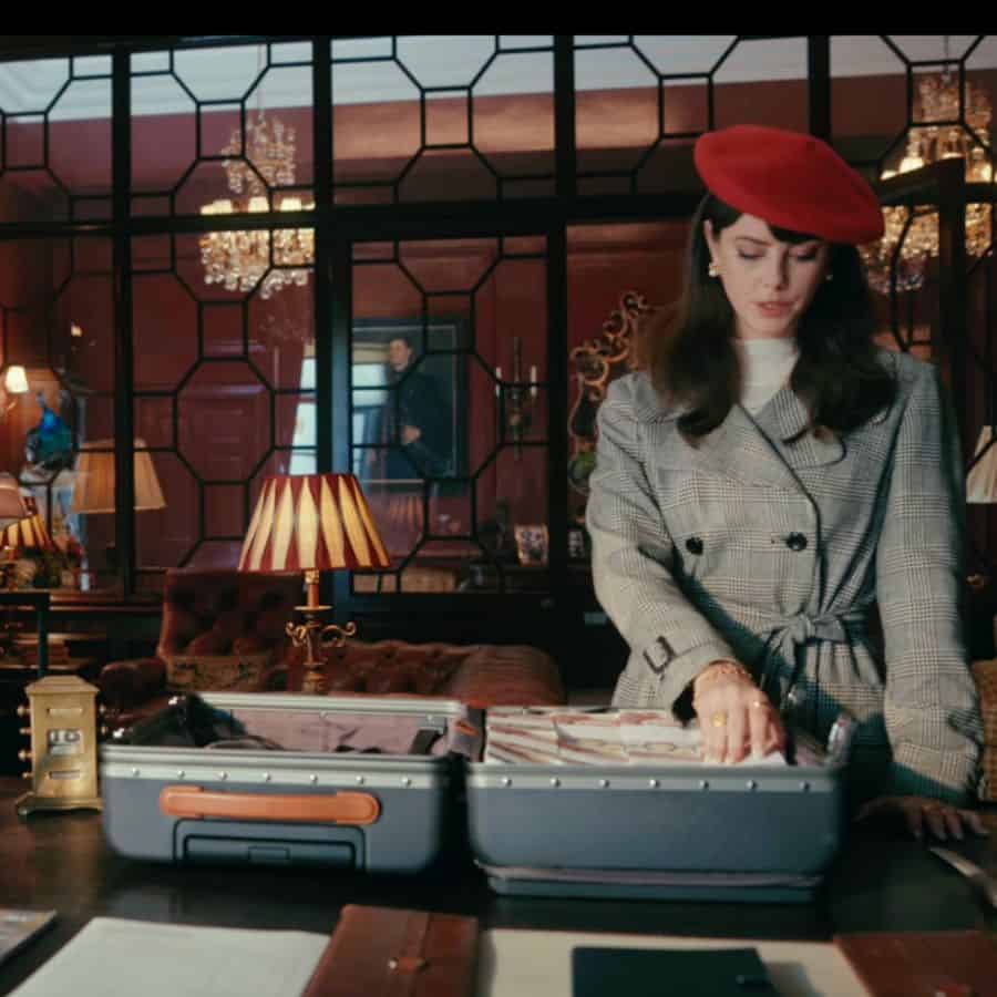 A youngish woman wearing a red beret and grey tweed coat is in a fancy room with her hand on a piece of open luggage. Inside the bag is piles of British pounds. Beyond her the room has portraits on maroon colored walls and chandeliers and other decorative items that suggest the home is old.
