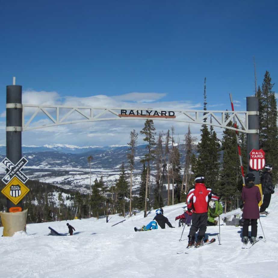 A sign marking the Railyard terrain park area at Winter Park ski resort with a group of people waiting at the top for their turn.