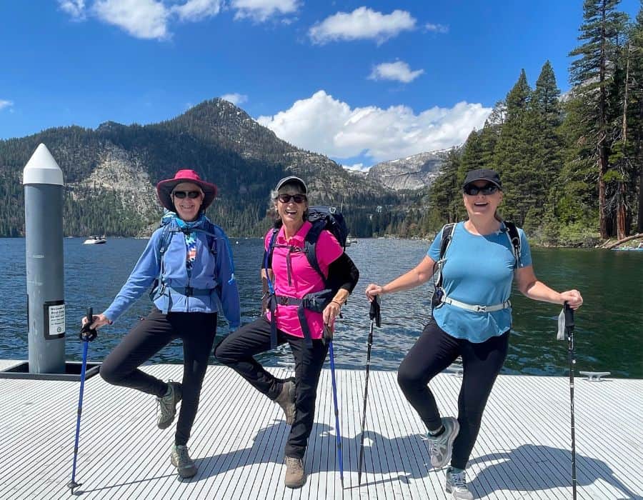 Three women hikers hold hiking poles and stand in tree pose, with a foot at their knee. They are at Lake Tahoe, California.